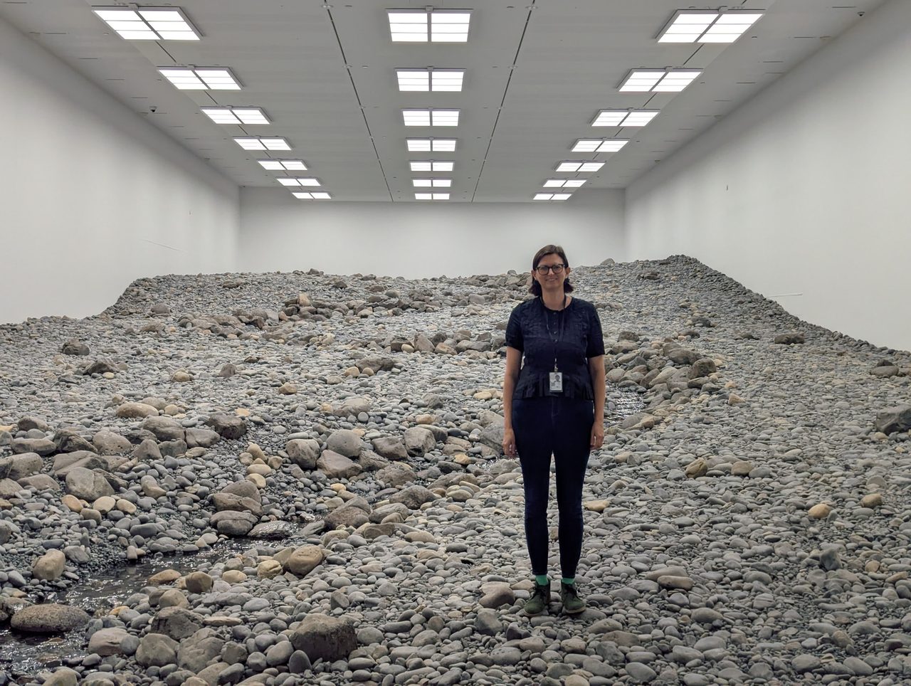 Wide view of the Riverbed gallery: rock-covered floor running to the back wall under rows of ceiling lights, with Megan Franks standing mid-frame.
