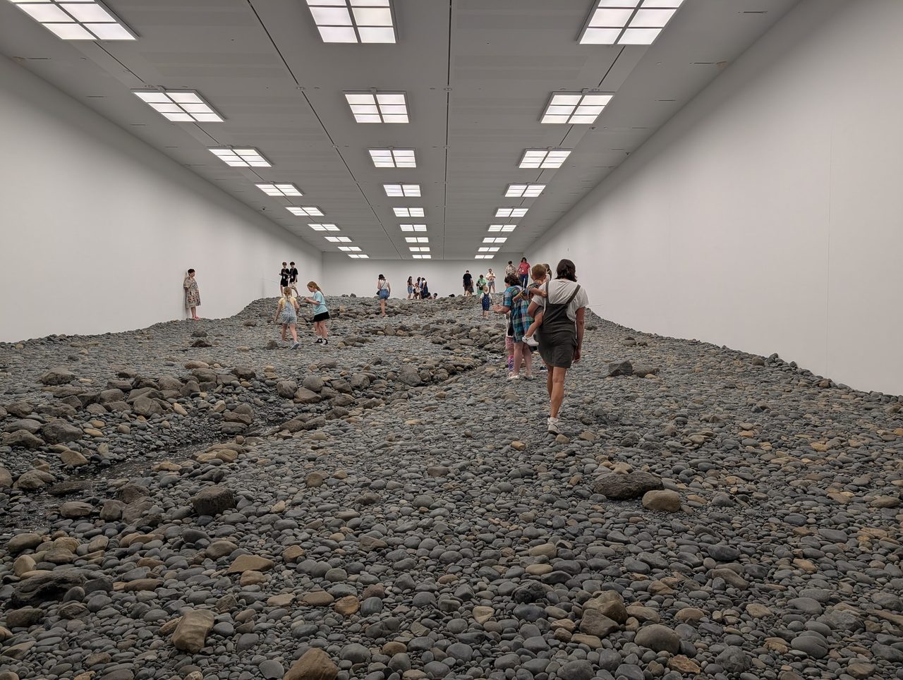 Wide view down the Riverbed gallery, visitors walking across the rocks between white walls under a grid of ceiling lights.