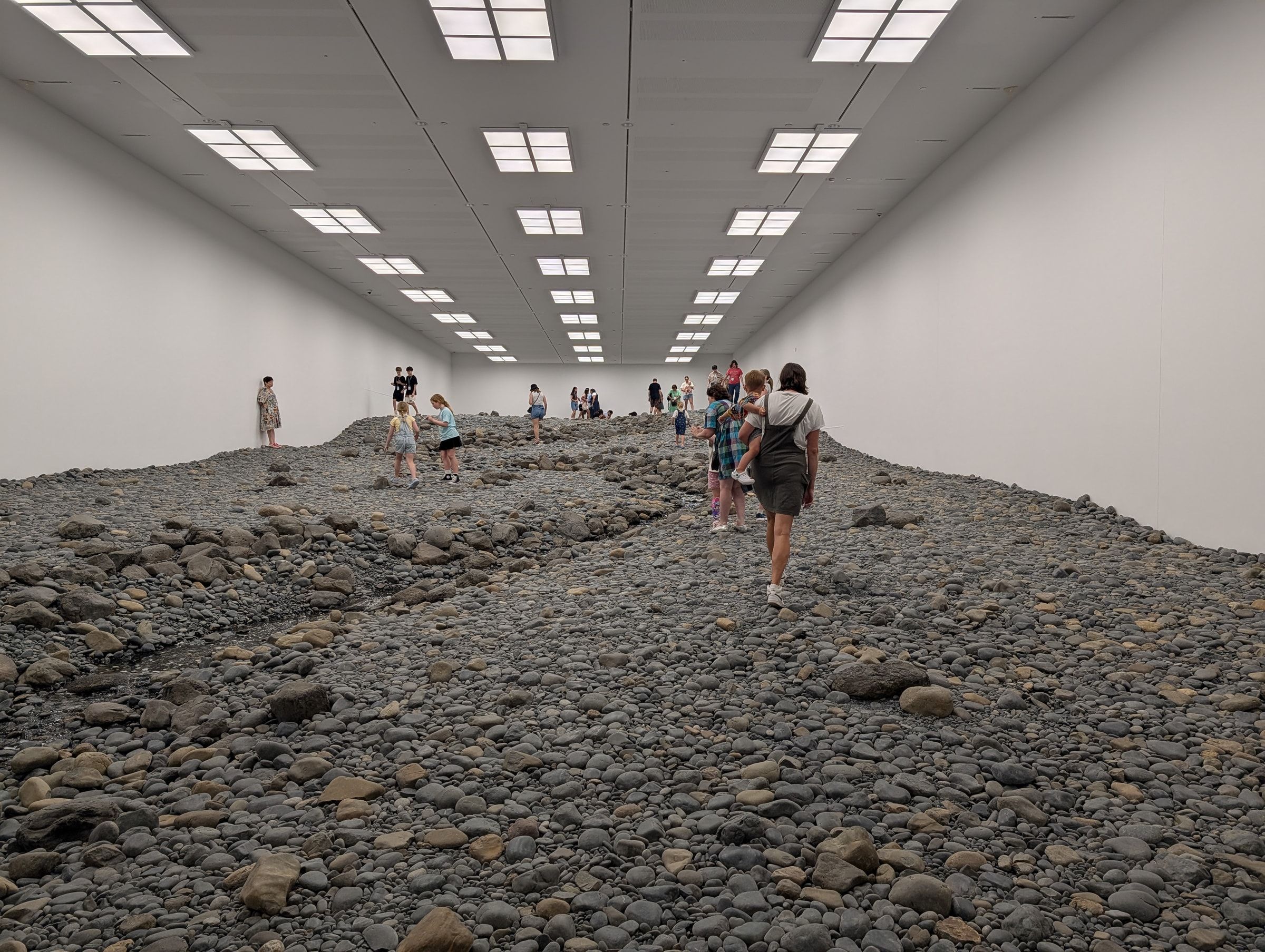 Wide view down the Riverbed gallery, visitors walking across the rocks between white walls under a grid of ceiling lights.