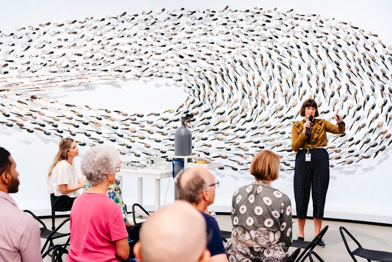 Megan Franks speaking with a microphone to a seated audience in front of a large swirling wall work made of feathered forms, during an APT9 talk.