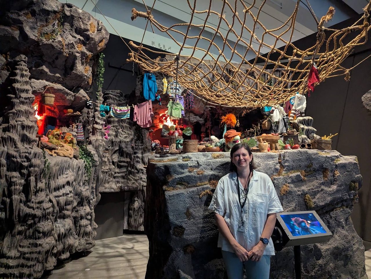 Megan Franks standing in front of the reconstructed Fraggle Rock set: rock caves, a rope net overhead, a washing line of small clothes and puppets on the ledges.