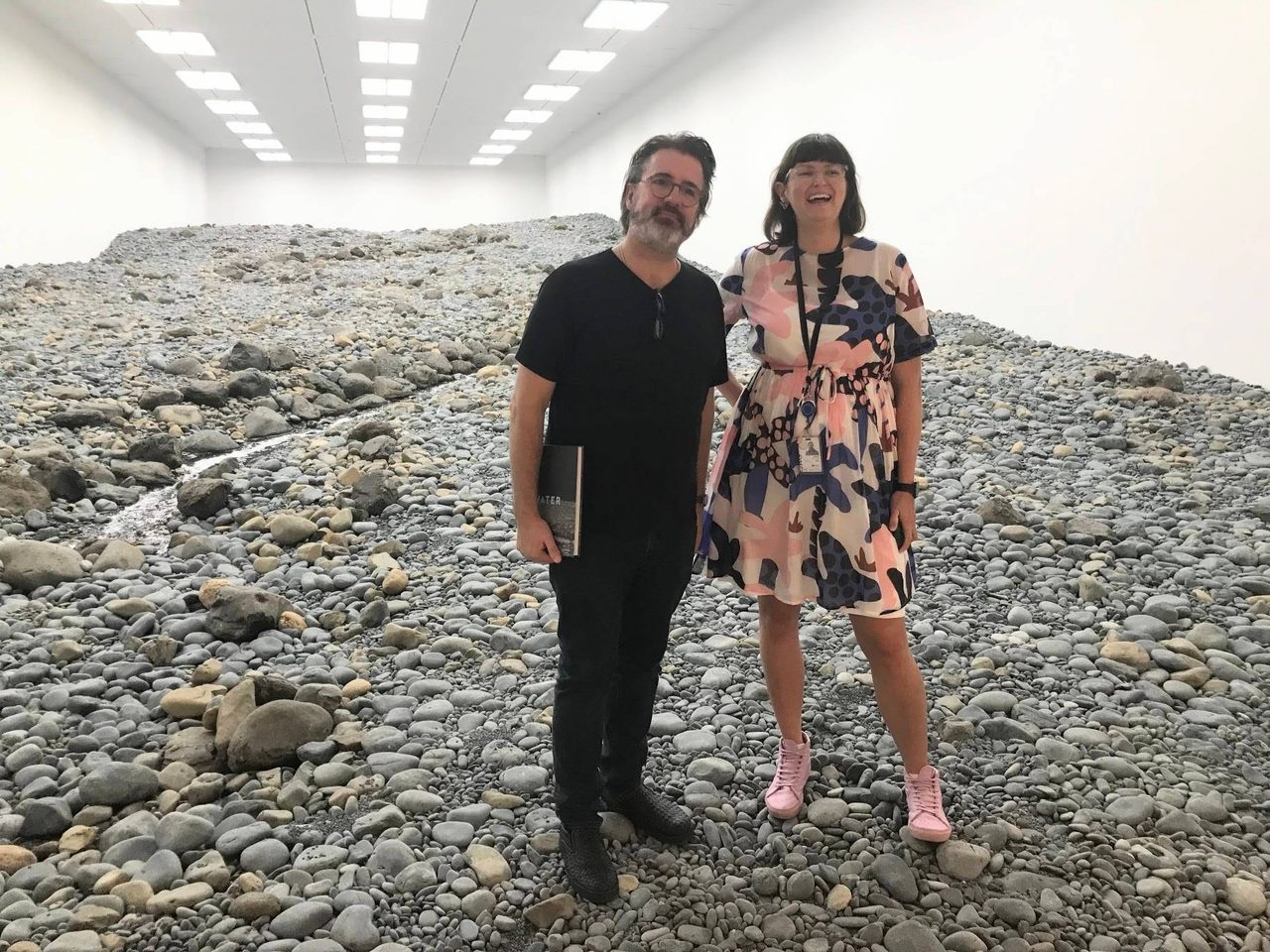 Wide view of the Riverbed gallery in 2019: a sloping field of grey river stones with a stream running down the left, white walls and rows of ceiling lights above, and Megan Franks standing right of centre beside the artist.